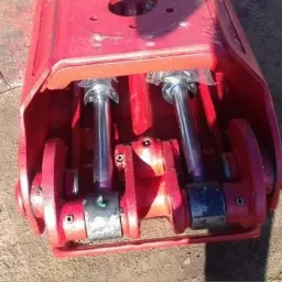 Czerwony element maszyny z dwoma chromowanymi cylindrami hydraulicznymi, widoczne ślady użytkowania i otarcia farby.