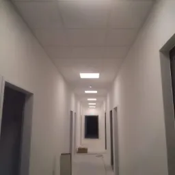 Długi, biały korytarz biurowy z podwieszanym sufitem kasetonowym i równomiernie rozmieszczonymi, wpuszczonymi lampami LED. Widoczne białe ściany, szare obramowania drzwi i okien oraz kartony...