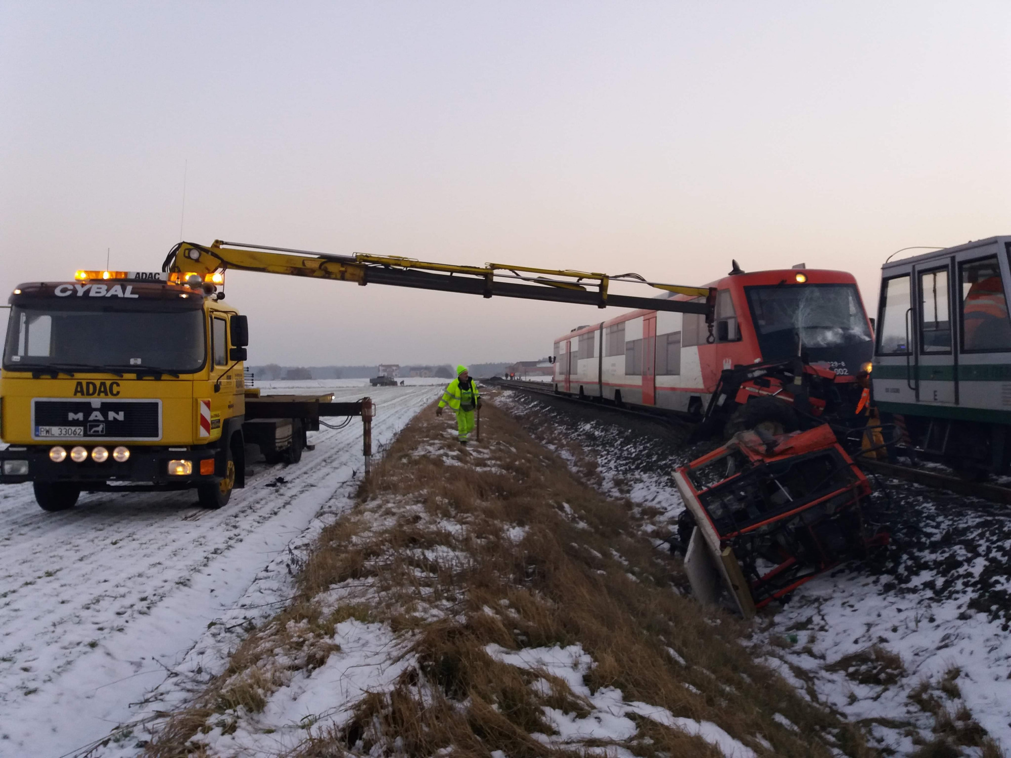 Żółta laweta ADAC z podniesionym ramieniem dźwigu przy wykolejonym pociągu i przewróconym ciągniku w zimowym krajobrazie, pracownik w odblaskowej kamizelce nadzoruje akcję.