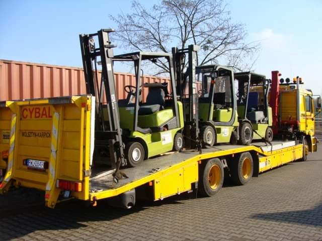 Żółta laweta z trzema wózkami widłowymi w kolorze oliwkowym, zabezpieczonymi pasami transportowymi, zaparkowana na kostce brukowej w słoneczny dzień.