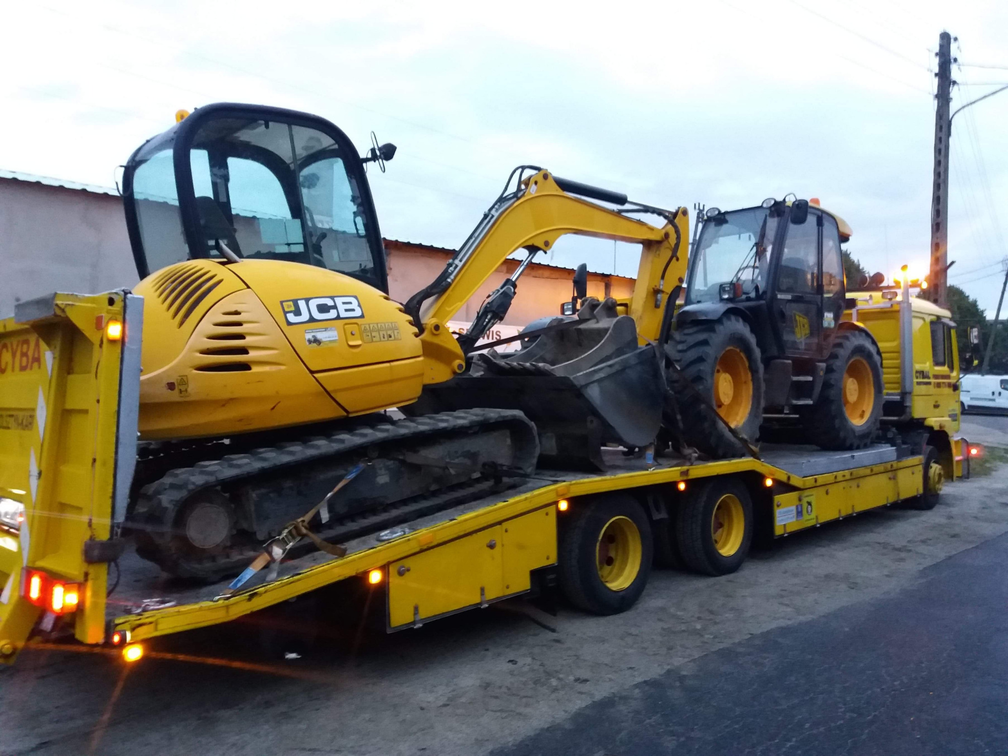 Żółta laweta z dwoma maszynami budowlanymi JCB: minikoparką na gąsienicach i koparko-ładowarką, zabezpieczonymi pasami transportowymi.