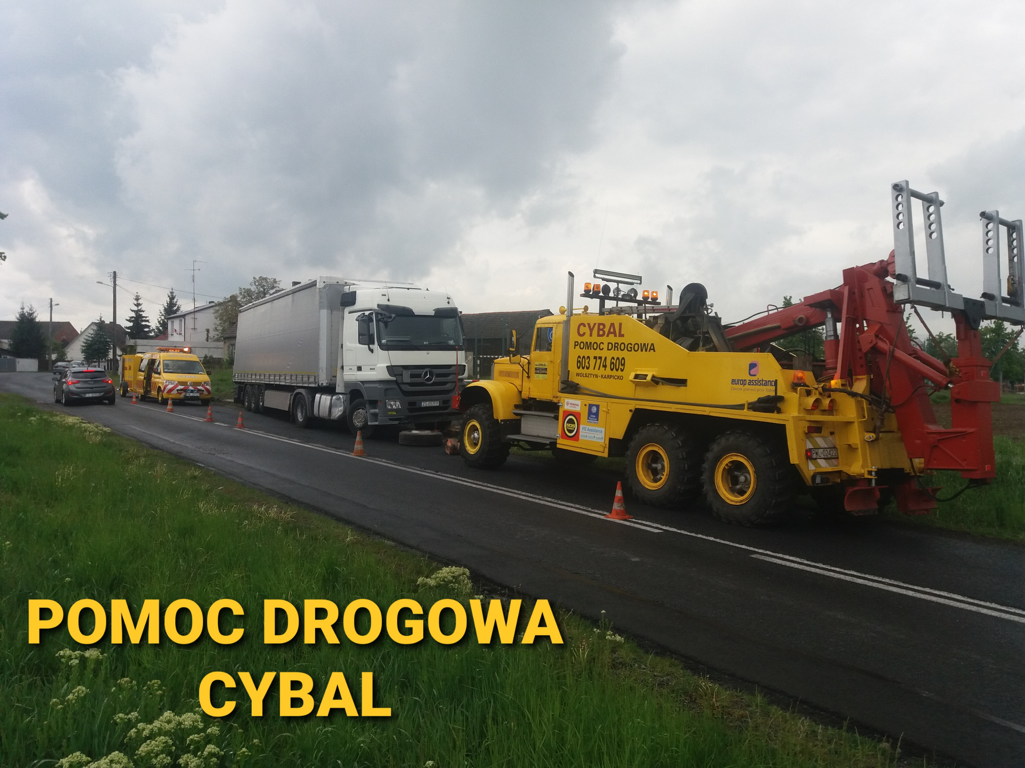 Żółta laweta firmy CYBAL z podnośnikiem holuje białą ciężarówkę na drodze, w tle stoi żółty bus pomocy drogowej i zaparkowany samochód osobowy.