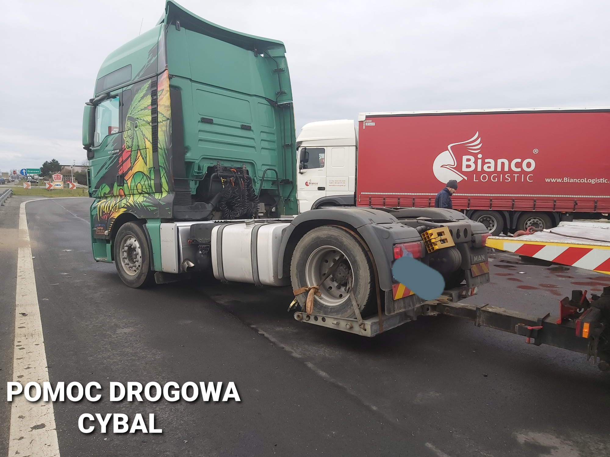 Zielony ciągnik siodłowy z grafiką indiańskiego motywu na kabinie, podpięty do lawety na autostradzie, w tle czerwona naczepa Bianco Logistic.