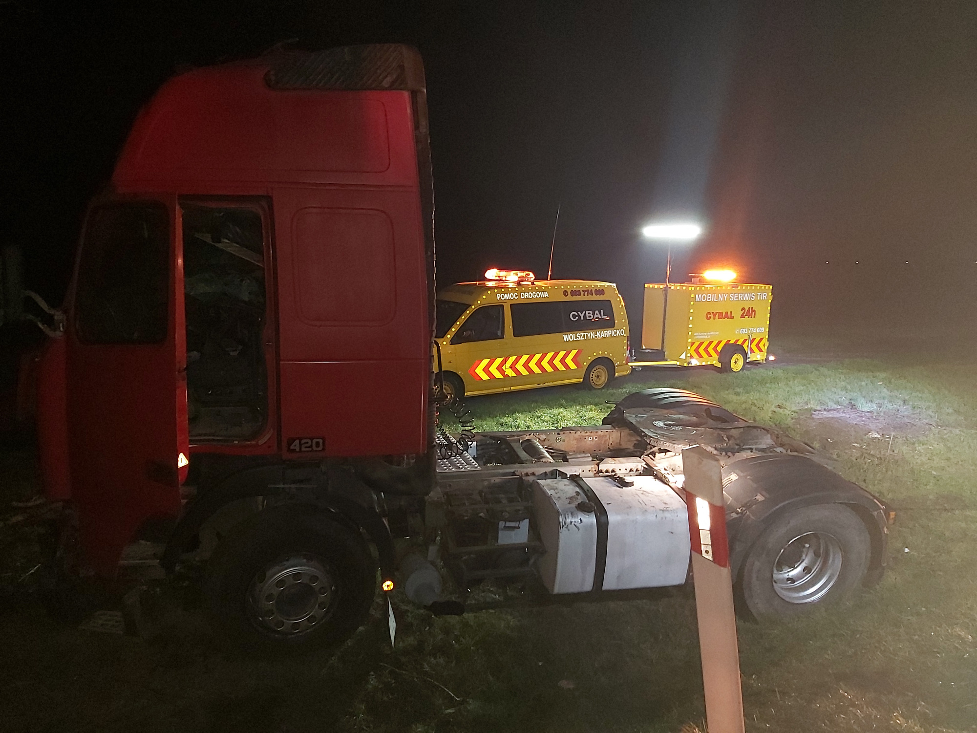 Czerwony ciągnik siodłowy bez naczepy stoi na trawie w nocy, obok żółty pojazd pomocy drogowej z przyczepą oświetlony światłami awaryjnymi i reflektorem.