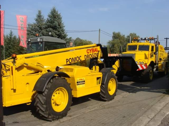 Żółty pojazd pomocy drogowej marki Cybal z podnośnikiem teleskopowym i ciężarówka holownicza w tle, widoczne detale opon i konstrukcji.