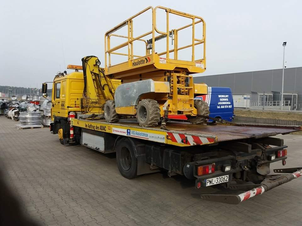 Żółta laweta z platformą transportową przewożąca podnośnik koszowy Haulotte, widoczny napis 'Im Auftrag des ADAC' oraz części samochodowe w tle.