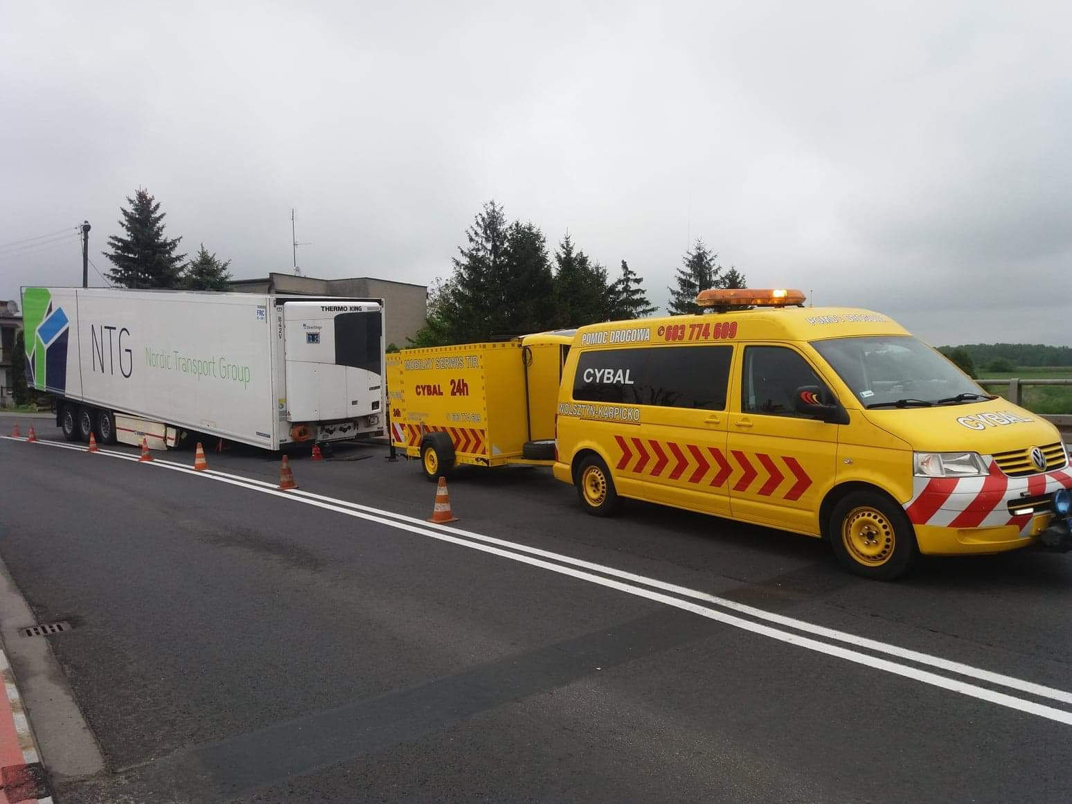 Żółty samochód pomocy drogowej firmy CYBAL z podpiętą przyczepą zabezpiecza uszkodzoną naczepę chłodniczą firmy NTG na drodze, ustawione pachołki ostrzegawcze.