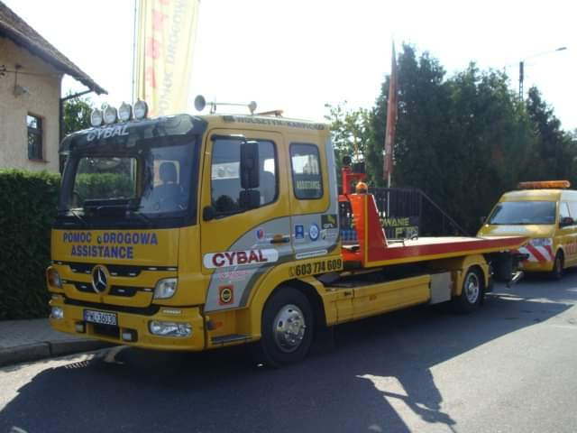 Żółta laweta Mercedes-Benz z napisem 'Pomoc Drogowa Assistance' i logo 'Cybal' na tle ulicy i budynku, z holowanym żółtym vanem.