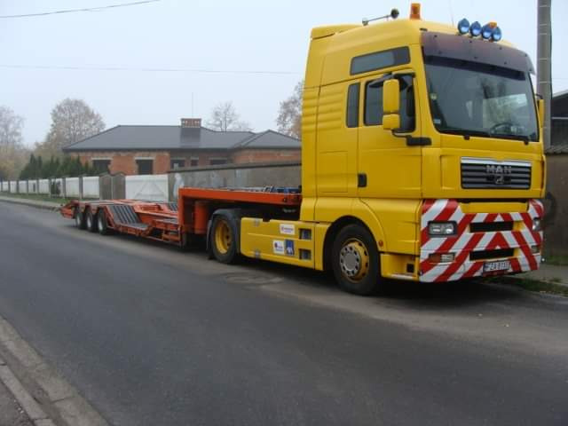 Żółta laweta MAN z niskopodwoziową naczepą na drodze, widoczny budynek w tle.