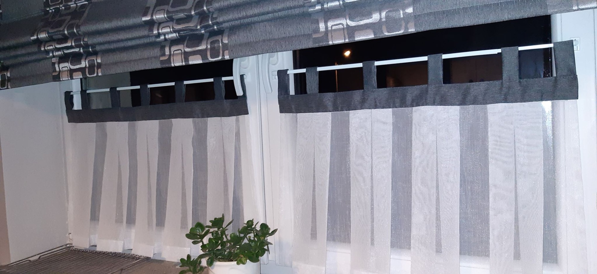 Dwa okna z szarymi roletami rzymskimi w geometryczny wzór i delikatnymi, białymi zasłonami z szarymi akcentami, zawieszonymi na białym karniszu z szarymi pętelkami, z rośliną doniczkową w dolnym rogu.