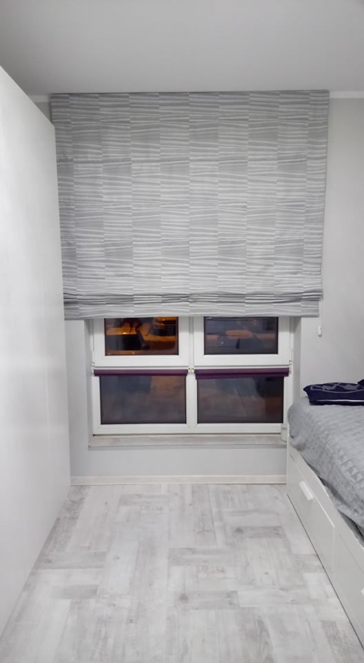 Zasłonięte okno w pokoju z szarą roletą rzymską w geometryczne wzory, widoczna część łóżka z szarą narzutą, biała podłoga i szafa.