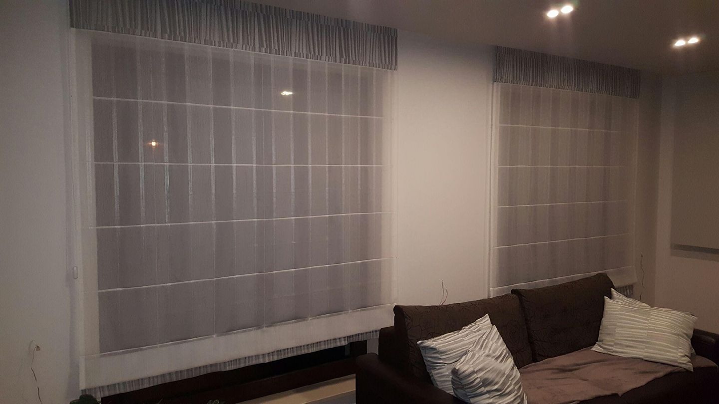 Dwa okna z szarymi, rzymskimi roletami z półprzezroczystej tkaniny w salonie, widoczne z brązowej sofy z poduszkami.