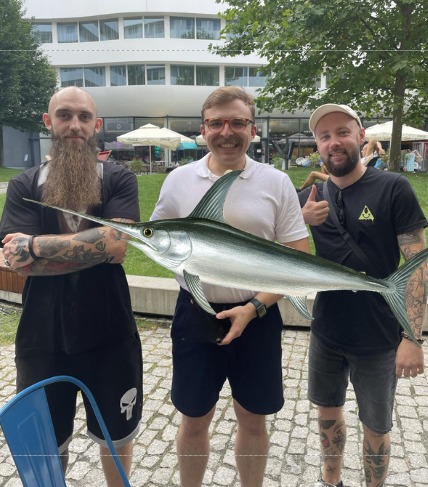 Trzech mężczyzn pozuje z cyfrowo dodaną rybą miecznik na tle budynku i zieleni.