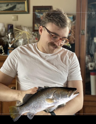 Mężczyzna w okularach i białej koszulce trzyma dużą rybę na tle mebli w domu.