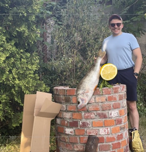 Mężczyzna w okularach przeciwsłonecznych i bordowej czapce pozuje z upolowaną rybą opartą o murowany grill z cegły, obok plaster cytryny i gałązka rozmarynu, w tle zielony żywopłot i kartonowe pudła.