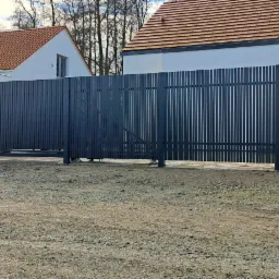 Nowoczesna, przesuwna brama wjazdowa z pionowych, metalowych profili w kolorze grafitowym, zintegrowana z furtką wyposażoną w wideodomofon i skrzynkę na listy, na tle domu z czerwoną dachówką.