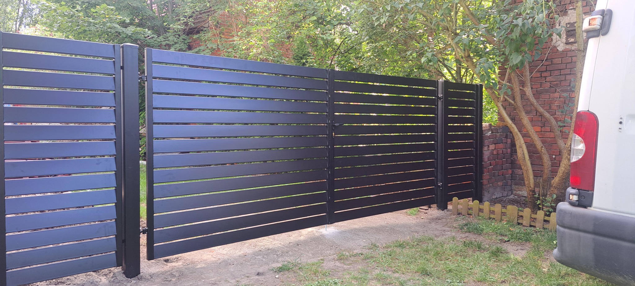 Nowoczesna, pozioma, czarna brama wjazdowa, widoczna od strony podwórza, z fragmentem białego samochodu dostawczego po prawej i ceglanym murem w tle.