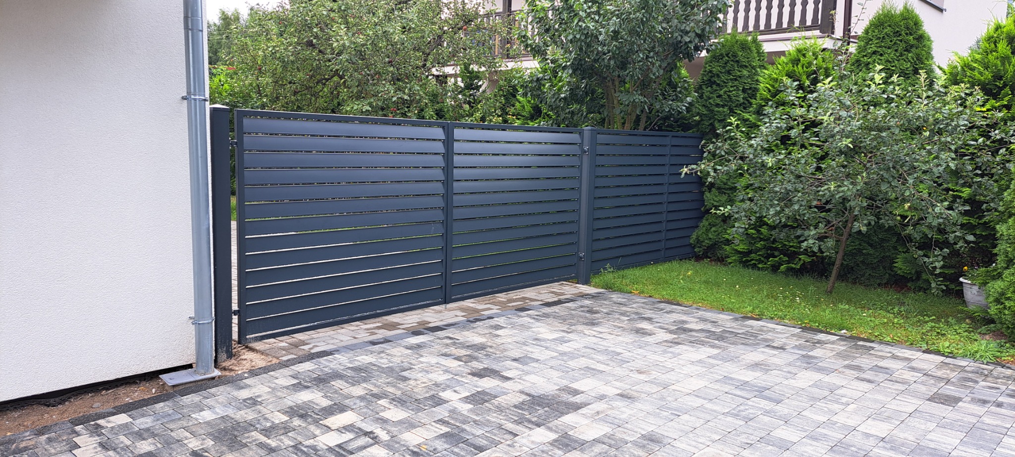 Nowoczesne, poziome, szare ogrodzenie metalowe z bramą wjazdową, widoczne z zewnątrz posesji, z kostką brukową na podjeździe i zielenią w tle.