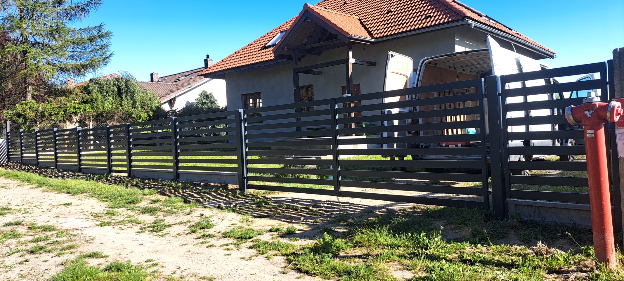 Nowoczesne, poziome ogrodzenie metalowe w kolorze grafitowym, z bramą wjazdową, na tle domu z czerwoną dachówką i zaparkowanego białego busa, widoczny czerwony hydrant.