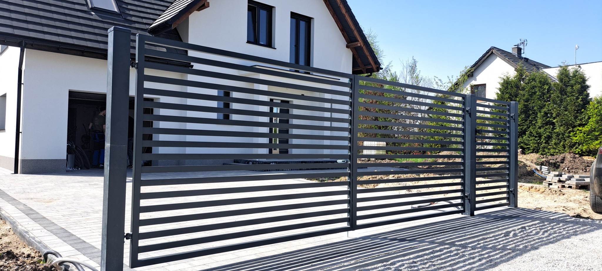 Nowoczesna, pozioma brama wjazdowa z metalowych profili, w kolorze antracytowym, zamontowana przed białym domem z czarnymi oknami i szarą kostką brukową, widoczne ślady prac ziemnych.