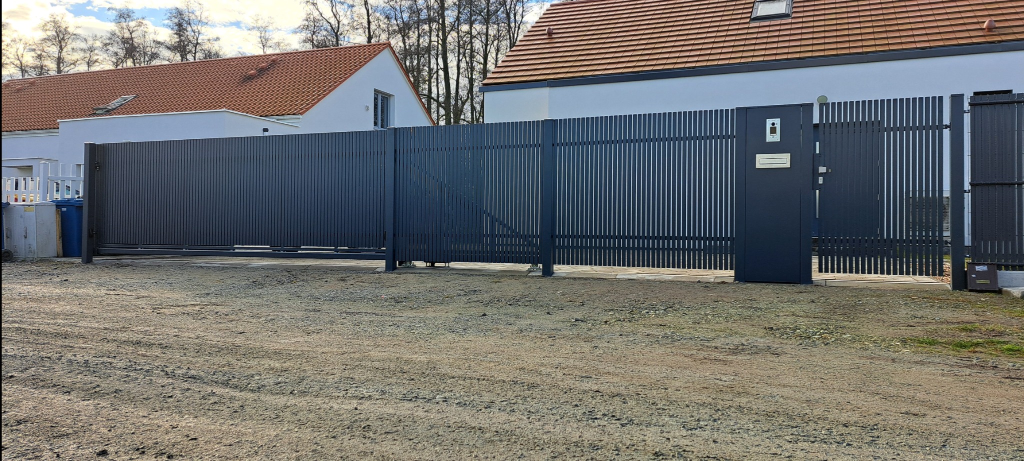 Nowoczesna, przesuwna brama wjazdowa z pionowych, metalowych profili w kolorze grafitowym, zintegrowana z furtką wyposażoną w wideodomofon i skrzynkę na listy, na tle domu z czerwoną dachówką.