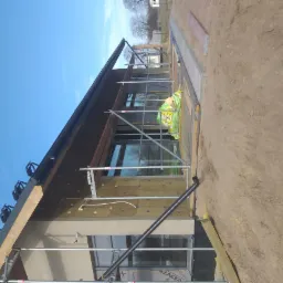 Elewacja budynku w trakcie ocieplania, widoczne rusztowania, izolacja z wełny mineralnej, okna oraz przygotowany teren wokół budynku.