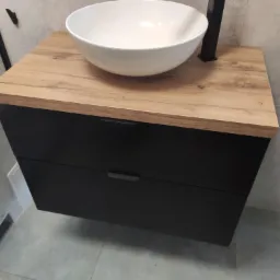 Umywalka nablatowa w kształcie misy, osadzona na szafce z dwoma szufladami o czarnych frontach i blacie imitującym jasne drewno, w nowoczesnej łazience z szarymi płytkami.