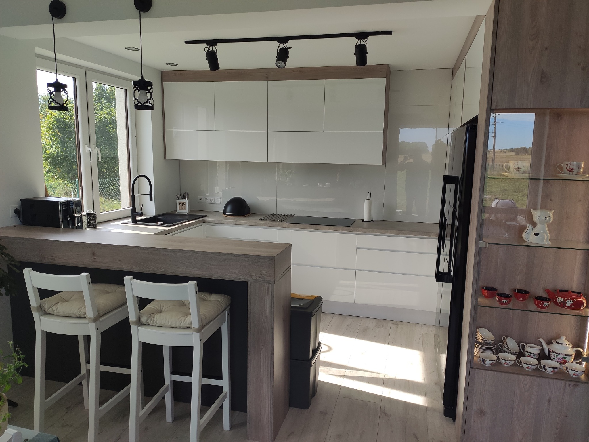 Jasna kuchnia w Krakowie z białymi szafkami, drewnianym blatem i dwoma białymi krzesłami barowymi przy wyspie. W tle widoczne okno z zielenią.