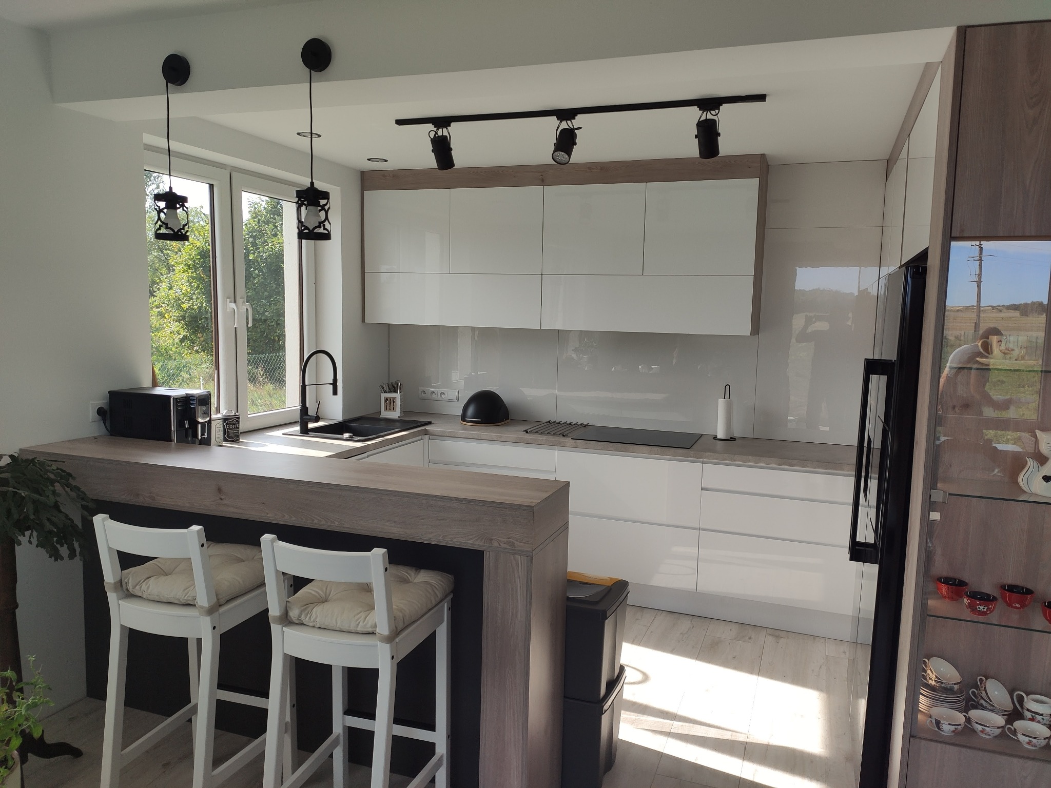 Jasna kuchnia z białymi frontami i drewnianym blatem, oświetlona wiszącymi lampami w stylu lampionów i reflektorami sufitowymi, z dwoma białymi krzesłami barowymi przy wyspie.