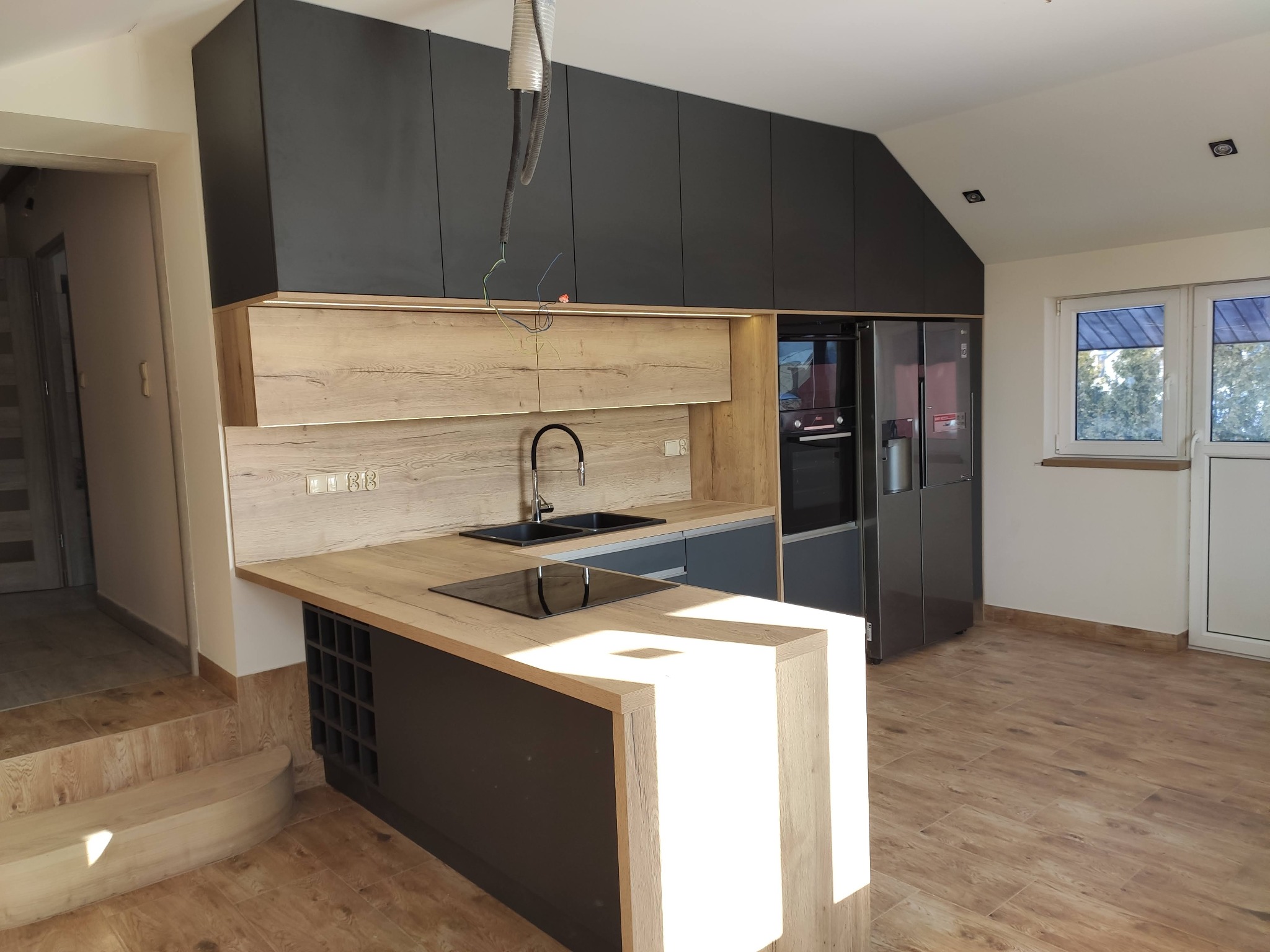 Nowoczesna kuchnia z wyspą, fronty w kolorze grafitowym i drewnopodobnym, widoczne niedokończone podłączenia instalacji nad szafkami.