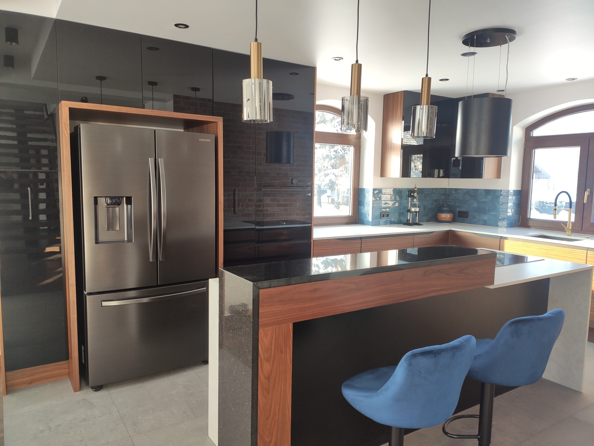 Nowoczesna kuchnia z czarnymi, lakierowanymi frontami szafek, wyspą z blatem z ciemnego kamienia i drewnianymi akcentami, oświetlona wiszącymi lampami z elementami szkła i złota, z widoczną lodówką...