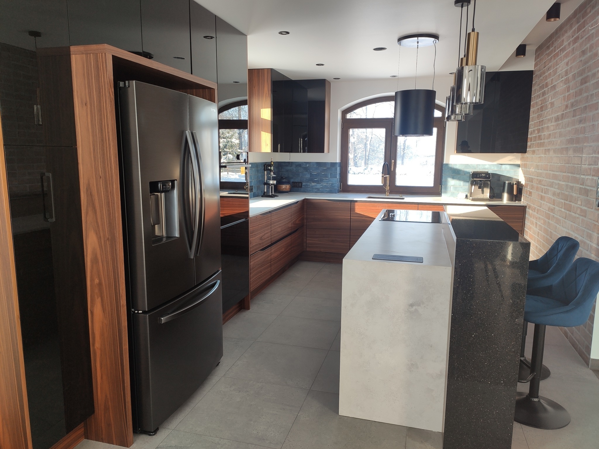 Nowoczesna kuchnia z drewnianymi frontami szafek, czarnymi elementami i wyspą z blatem imitującym beton, oświetlona wiszącymi lampami i światłem wpadającym przez łukowe okno.