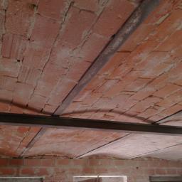 Sandblast - Ceglany łukowy strop wzmocniony stalowymi profilami, widok z dołu na tle surowych ścian budynku.