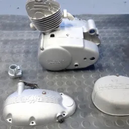 Elementy silnika motocyklowego Predom-Dezamet po renowacji, pokryte srebrzystą farbą, leżące na gumowej macie. Widoczny cylinder, karter i dwie pokrywy.