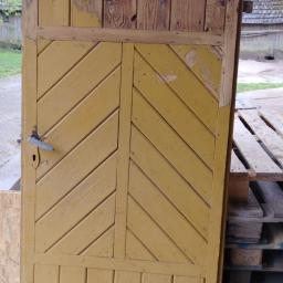 Sandblast - Stara, drewniana, żółtawa brama z widocznymi śladami zużycia i ubytkami farby, oparta o paletę. Górna część bramy z surowego drewna.