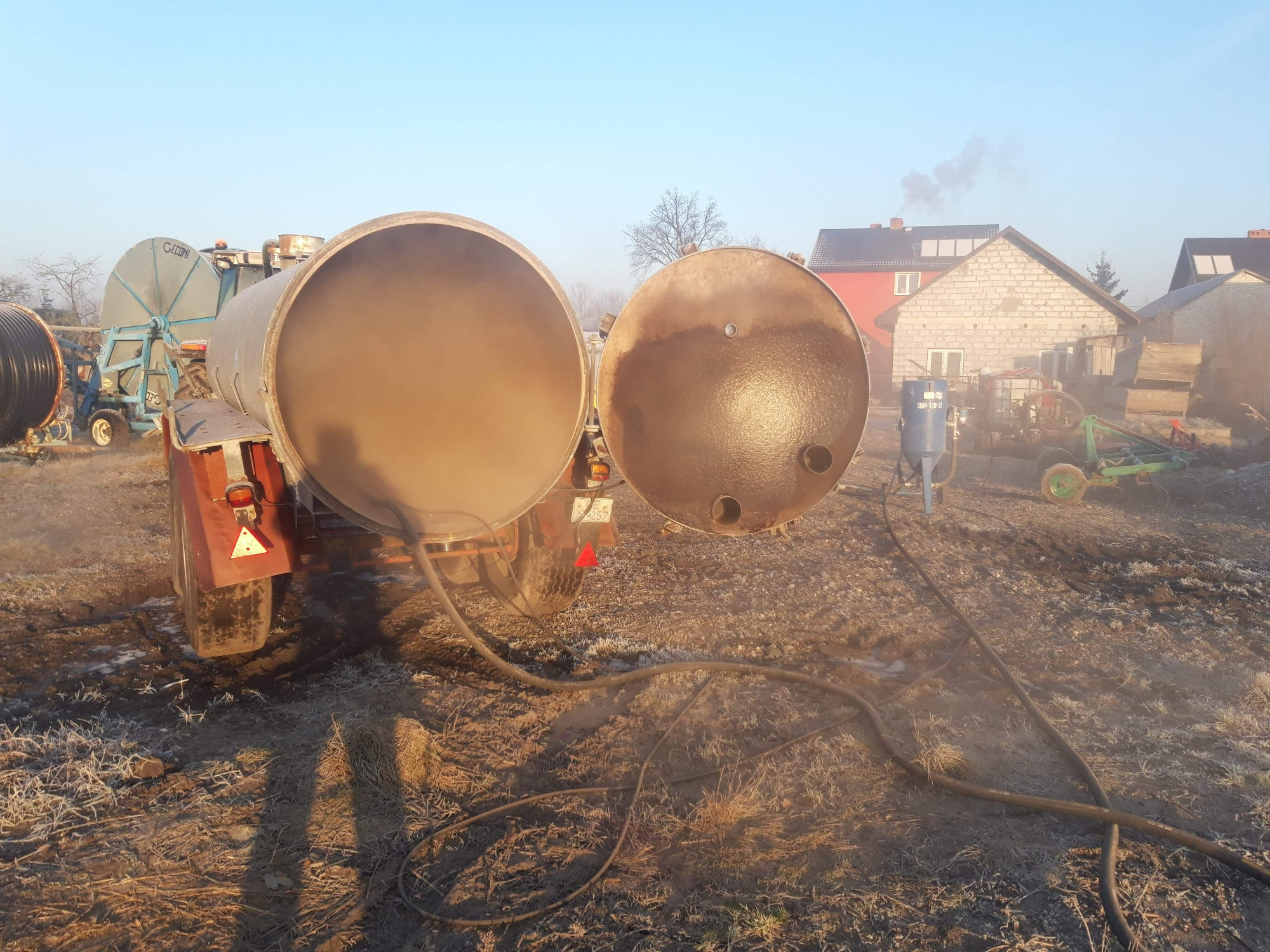 Otwarta cysterna na przyczepie, widoczne ślady czyszczenia strumieniowego, w tle budynki gospodarcze i sprzęt rolniczy, poranna mgła.
