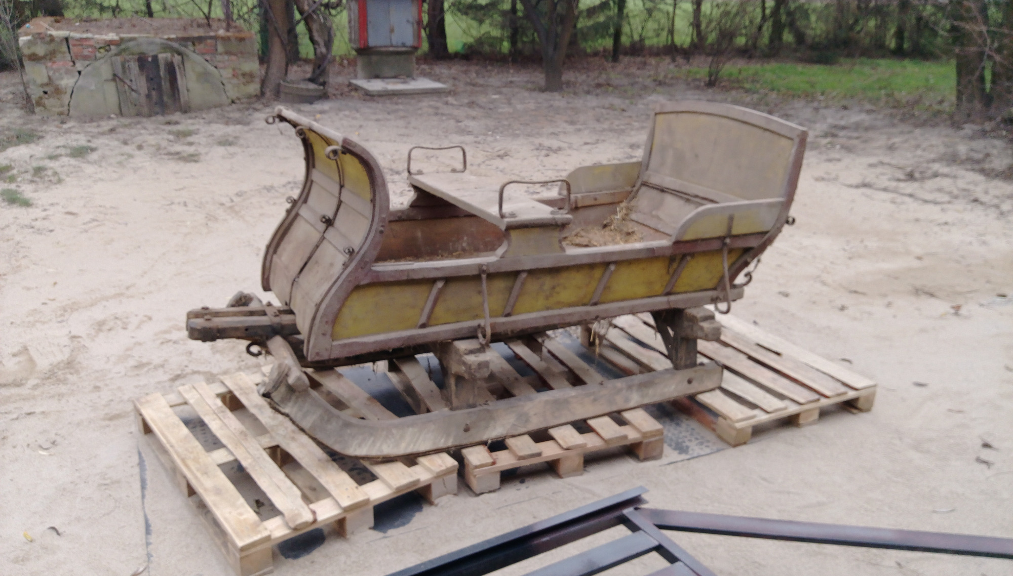 Stara, drewniana sanie na paletach, z widocznymi śladami rdzy i żółtą farbą, w tle ziemisty teren i zabudowa przypominająca piwniczkę.