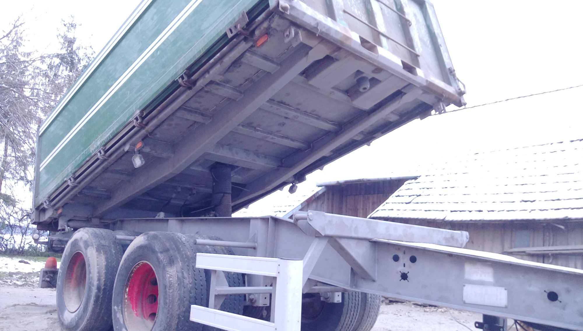Przyczepa wywrotka z podniesioną zieloną skrzynią ładunkową, widoczne czerwone felgi kół i konstrukcja nośna, w tle budynek gospodarczy ze śniegiem na dachu.