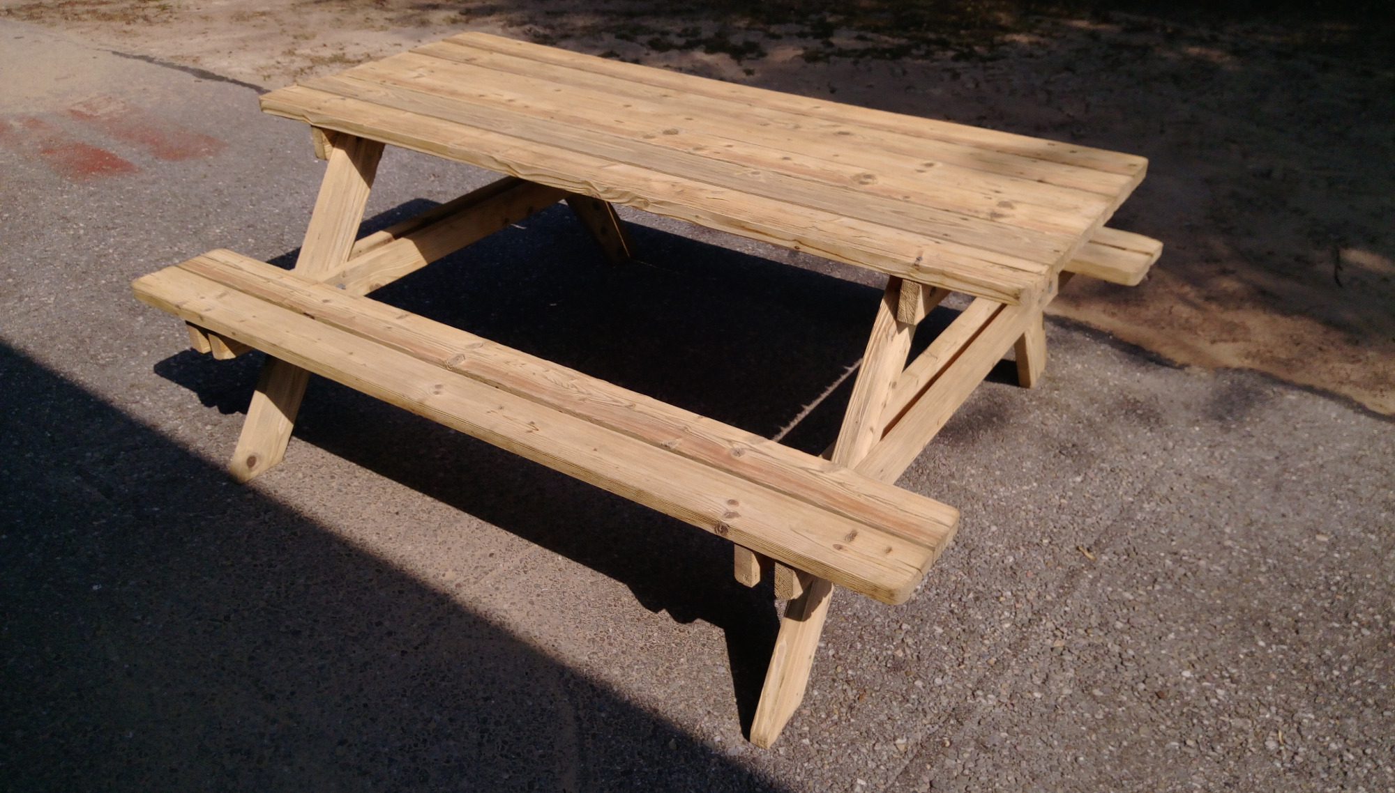 Nowy, surowy drewniany stół piknikowy z ławkami, stojący na szarym betonowym podłożu w słoneczny dzień.