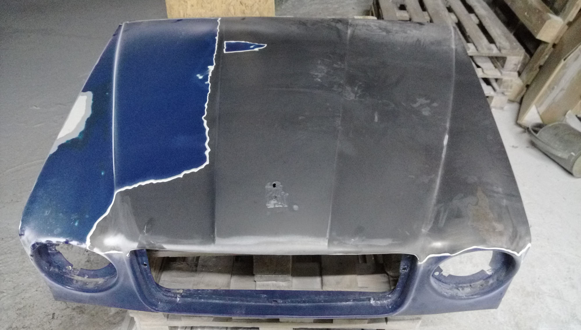 Częściowo odnowiona maska samochodu na palecie, z widocznymi fragmentami starej i nowej powłoki lakierniczej, przygotowana do dalszych prac.