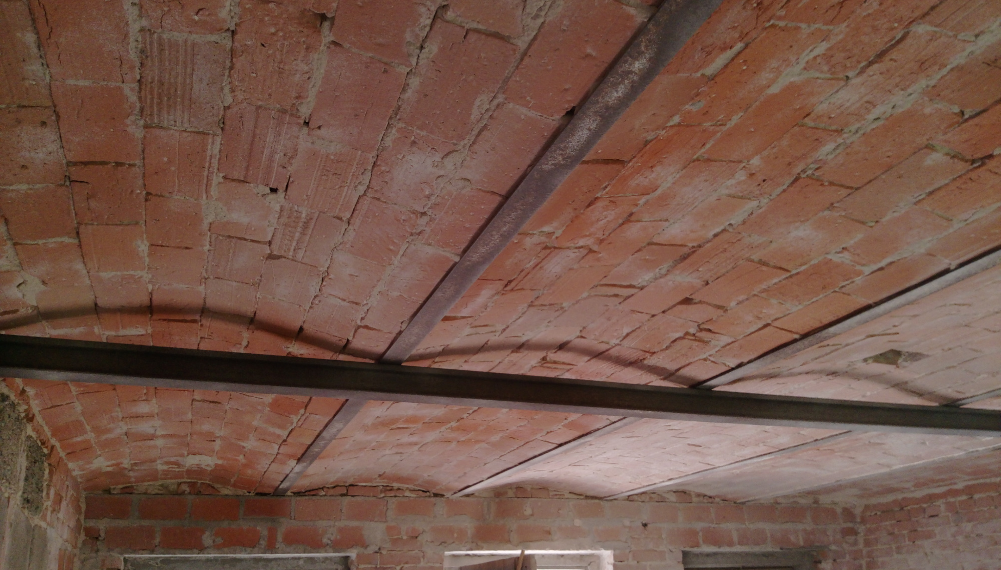 Ceglany łukowy strop wzmocniony stalowymi profilami, widok z dołu na tle surowych ścian budynku.