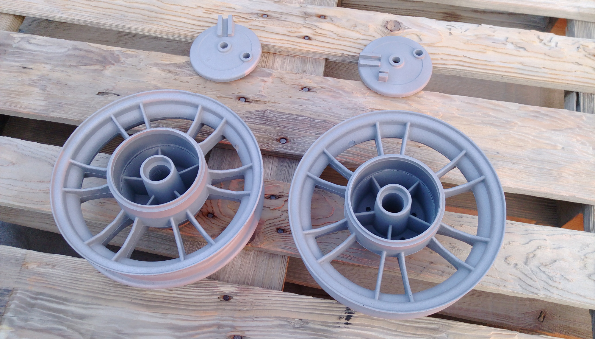 Dwa szare, metalowe koła i dwa okrągłe elementy, prawdopodobnie po procesie nałożenia powłoki, leżące na drewnianej palecie. Widoczne delikatne refleksy światła na powierzchni.