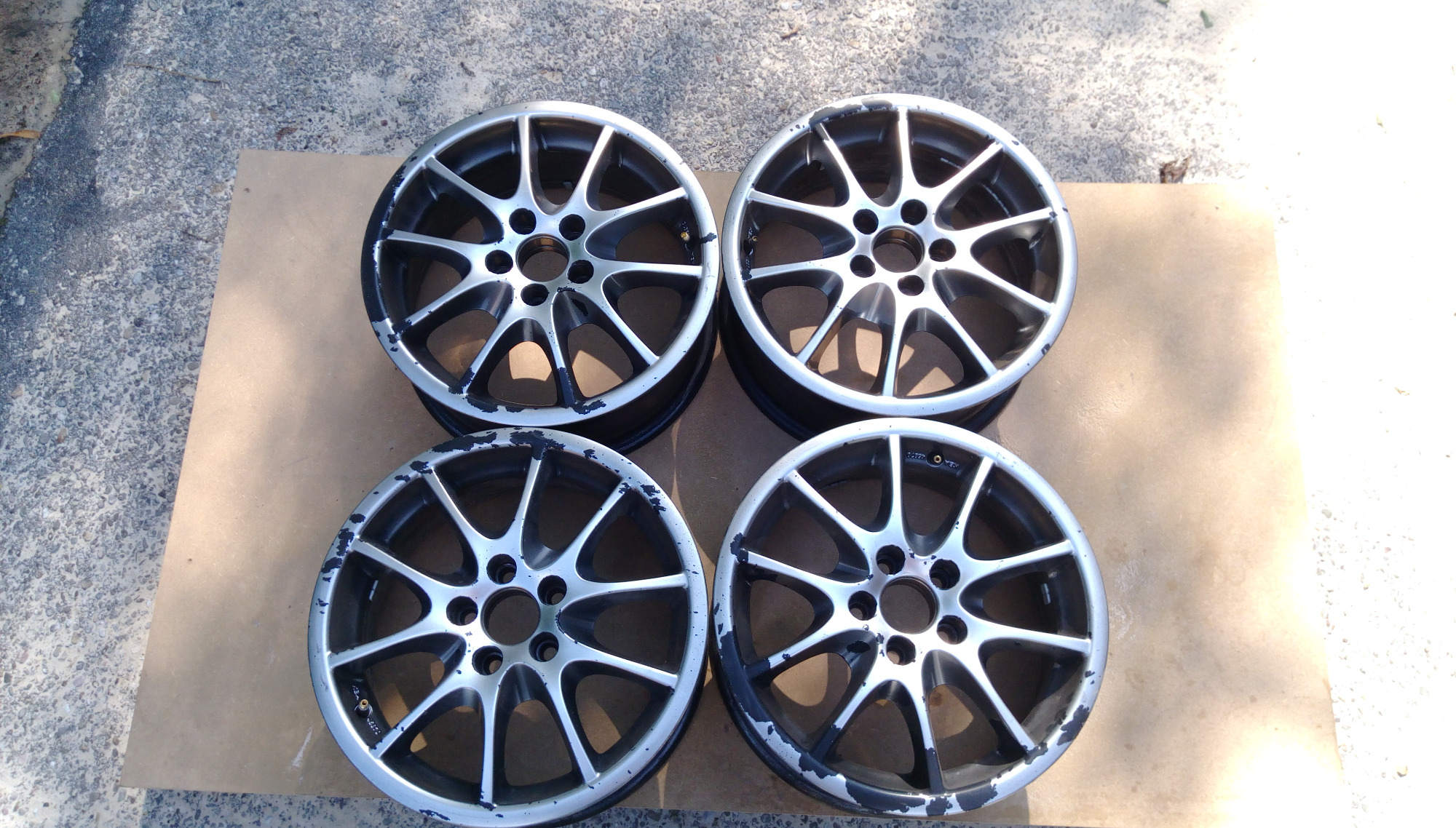Cztery aluminiowe felgi samochodowe z uszkodzoną powłoką lakierniczą, ułożone na kartonie na zewnątrz, przygotowane do renowacji.