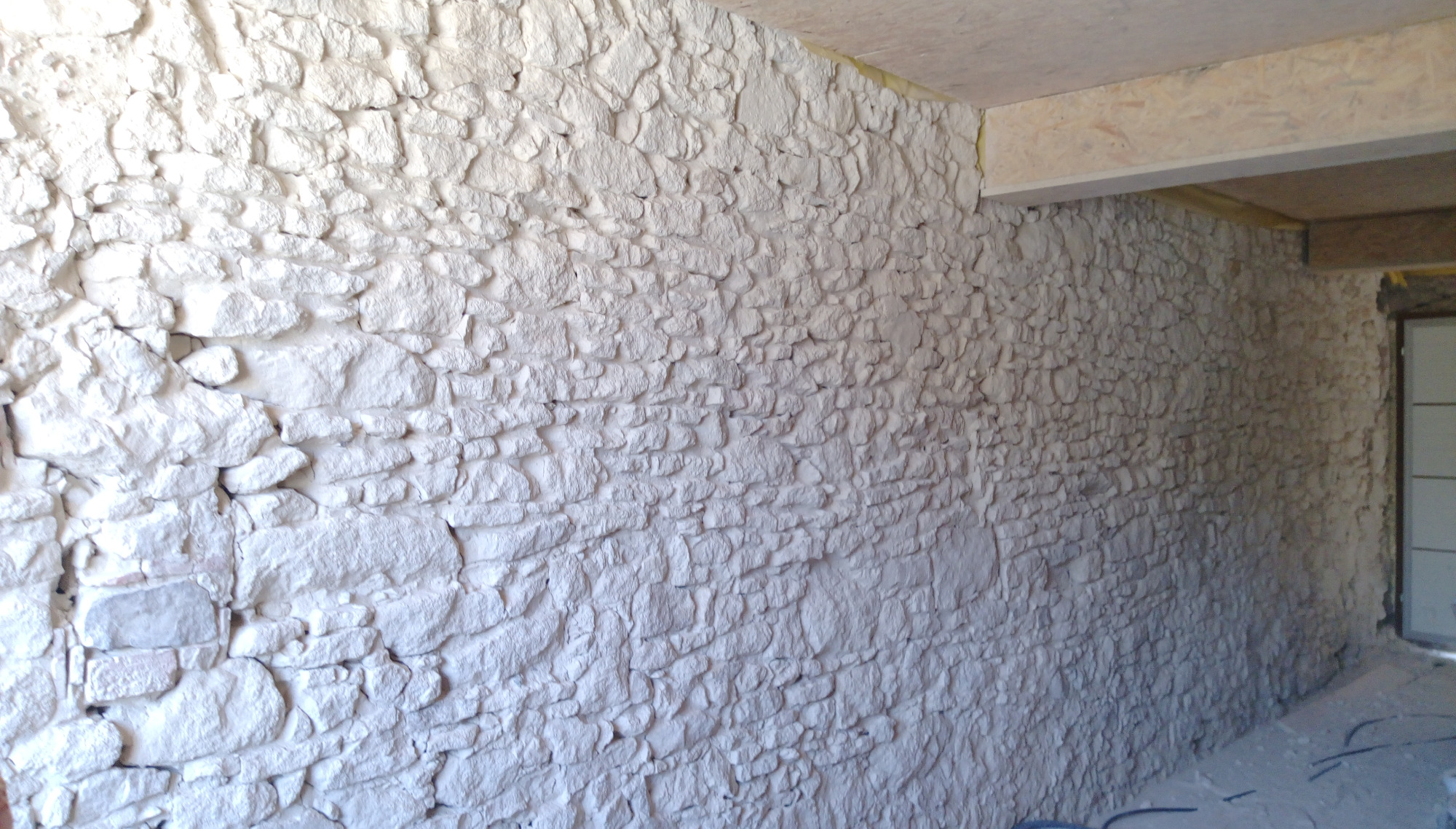 Wnętrze budynku z nierówną, białą ścianą imitującą kamień, widoczne fragmenty sufitu i drzwi w tle.