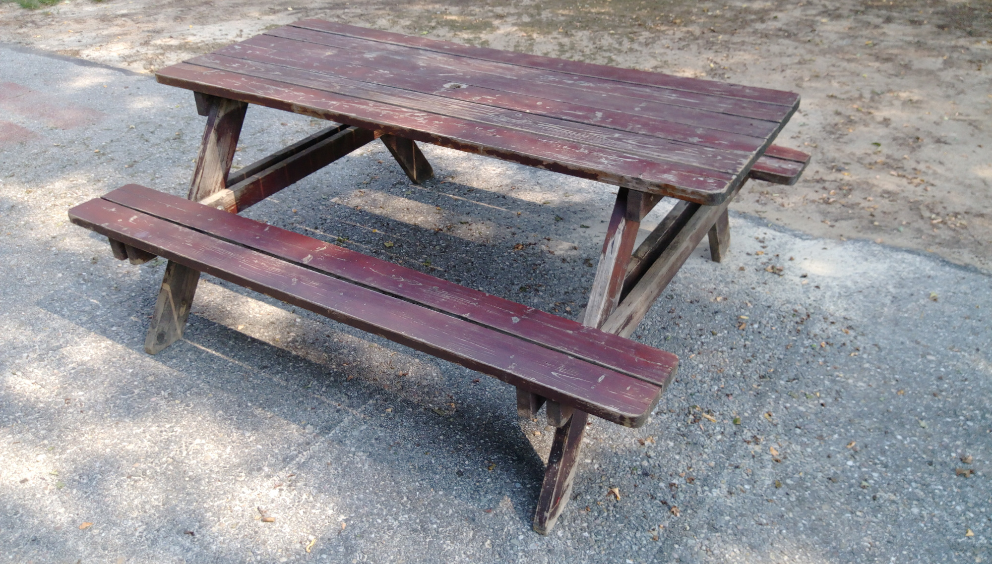 Drewniany stół piknikowy z ławkami, widoczne ślady użytkowania i odpryski farby, ustawiony na szarym podłożu z drobnych kamieni.