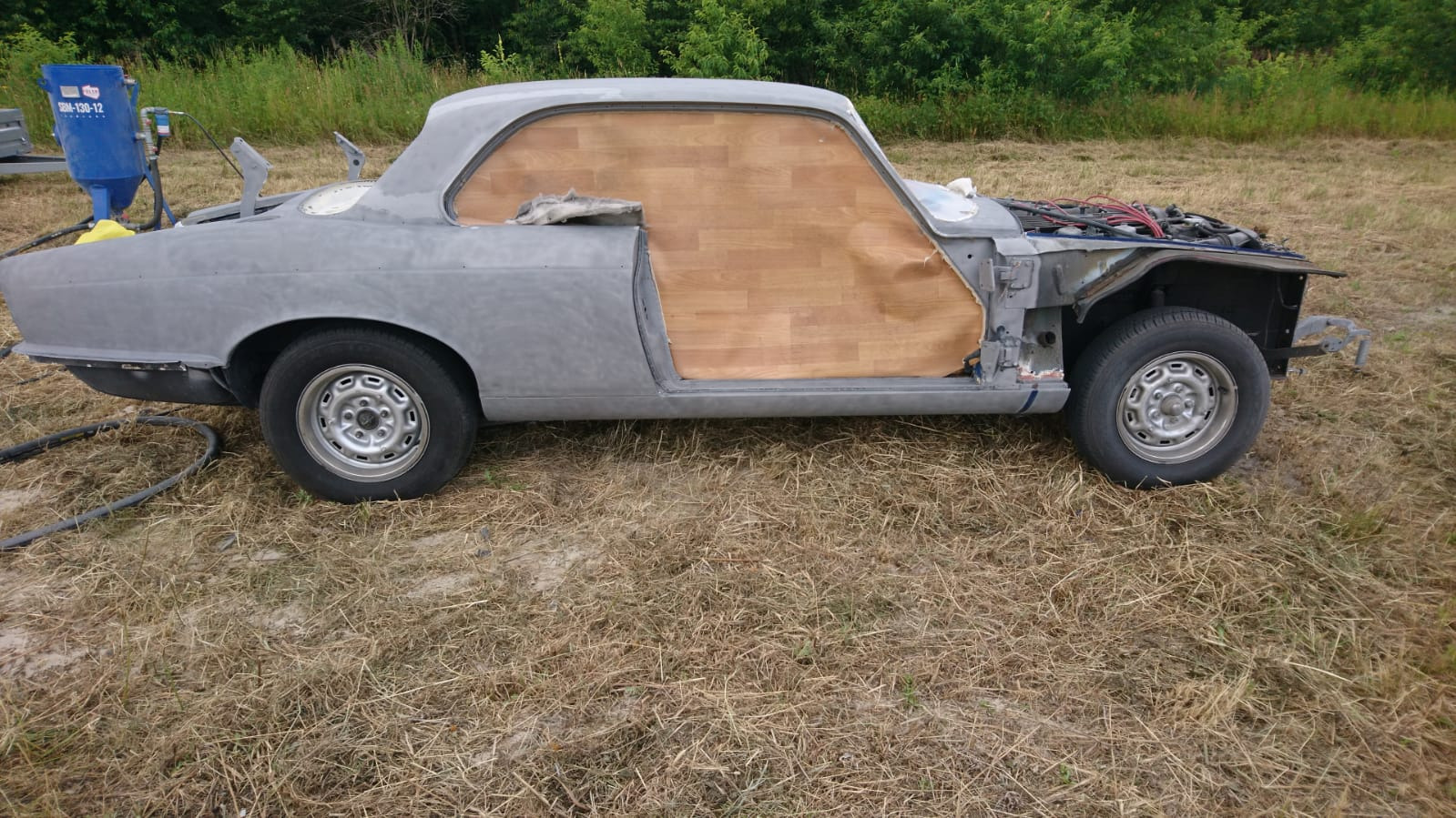 Klasyczny samochód podczas renowacji, z elementami karoserii po piaskowaniu i zabezpieczonymi drewnianymi panelami, widoczna maszyna do piaskowania w tle.