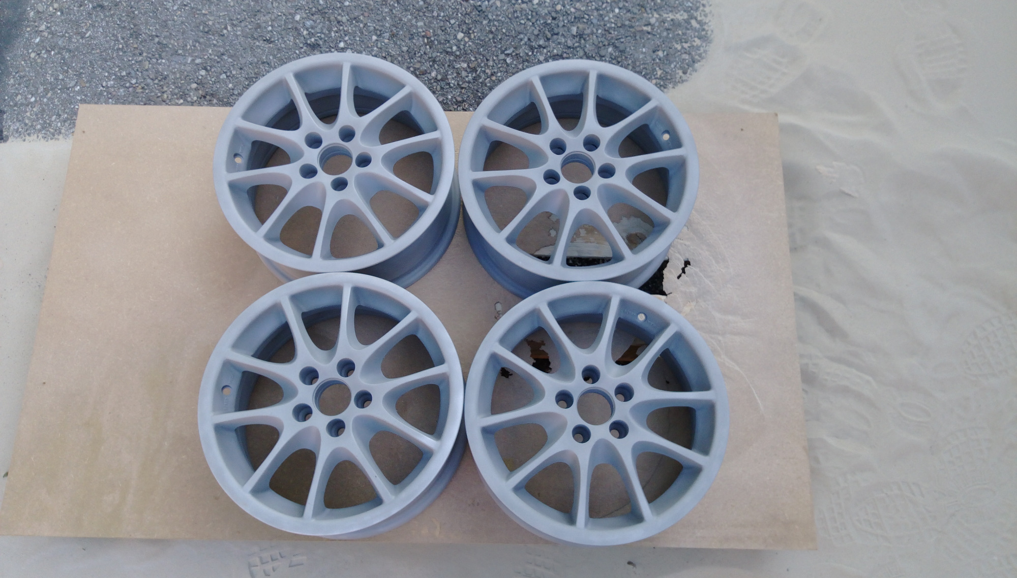 Cztery felgi samochodowe po obróbce, leżące na kartonie na zewnątrz, pokryte świeżą warstwą szarego podkładu.