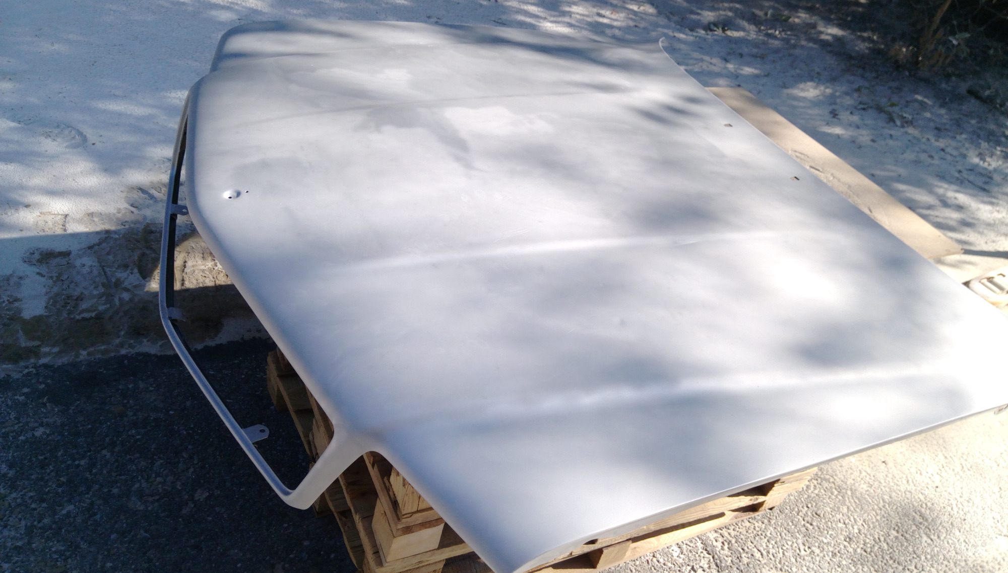 Element karoserii samochodu, prawdopodobnie maska, po wstępnej obróbce, leżący na palecie w plenerze, przygotowany do dalszych prac wykończeniowych.