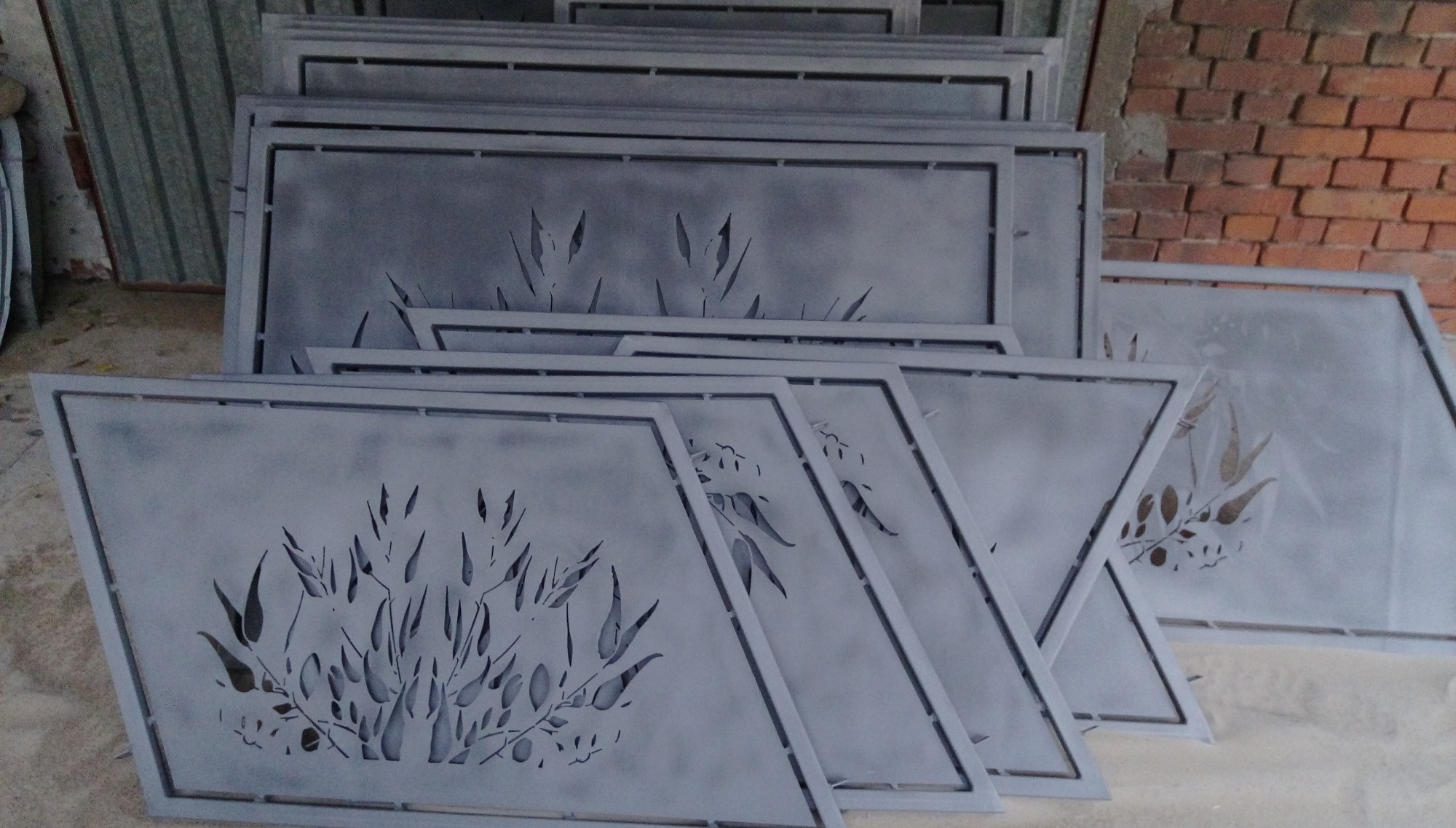 Stos metalowych paneli z wyciętym motywem roślinnym, po obróbce powierzchniowej, ustawionych pod ścianą z cegły.