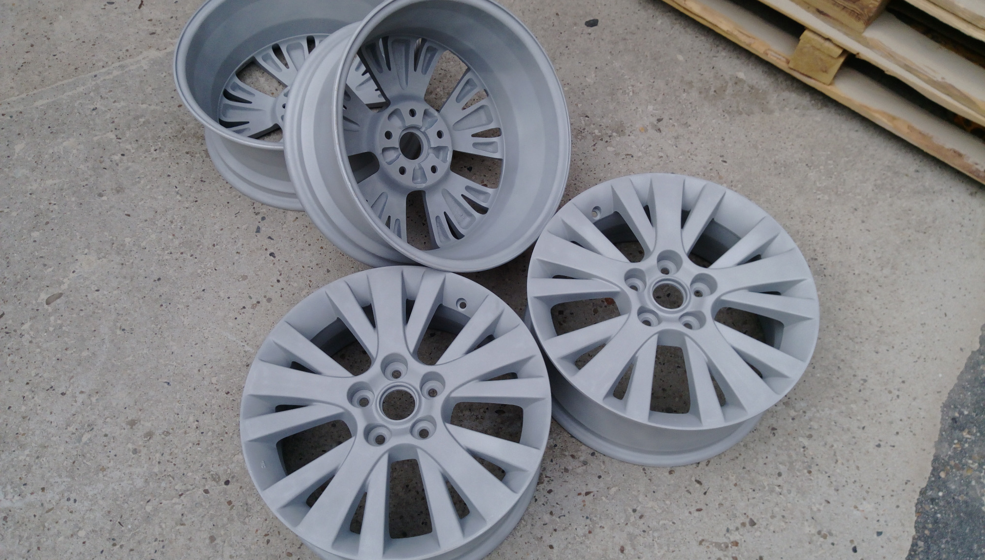Cztery felgi samochodowe w szarym kolorze, leżące na betonowej powierzchni, obok drewnianej palety, po procesie malowania.